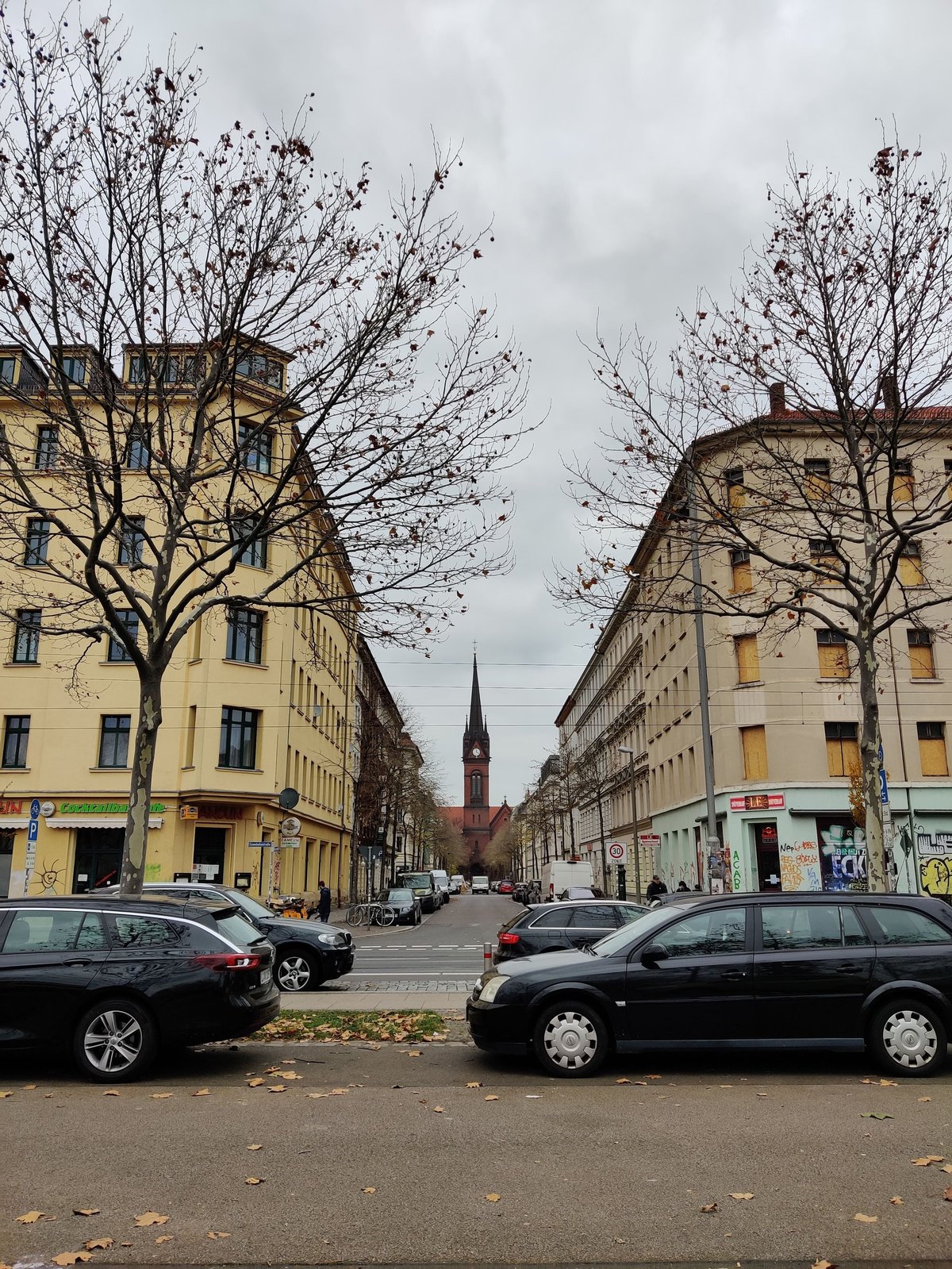 Leipziger Stadtstraße mit gelben Gründerzeit-Häusern, kahlen Bäumen, parkenden Autos und Kirchturm in der Ferne unter bedecktem Himmel